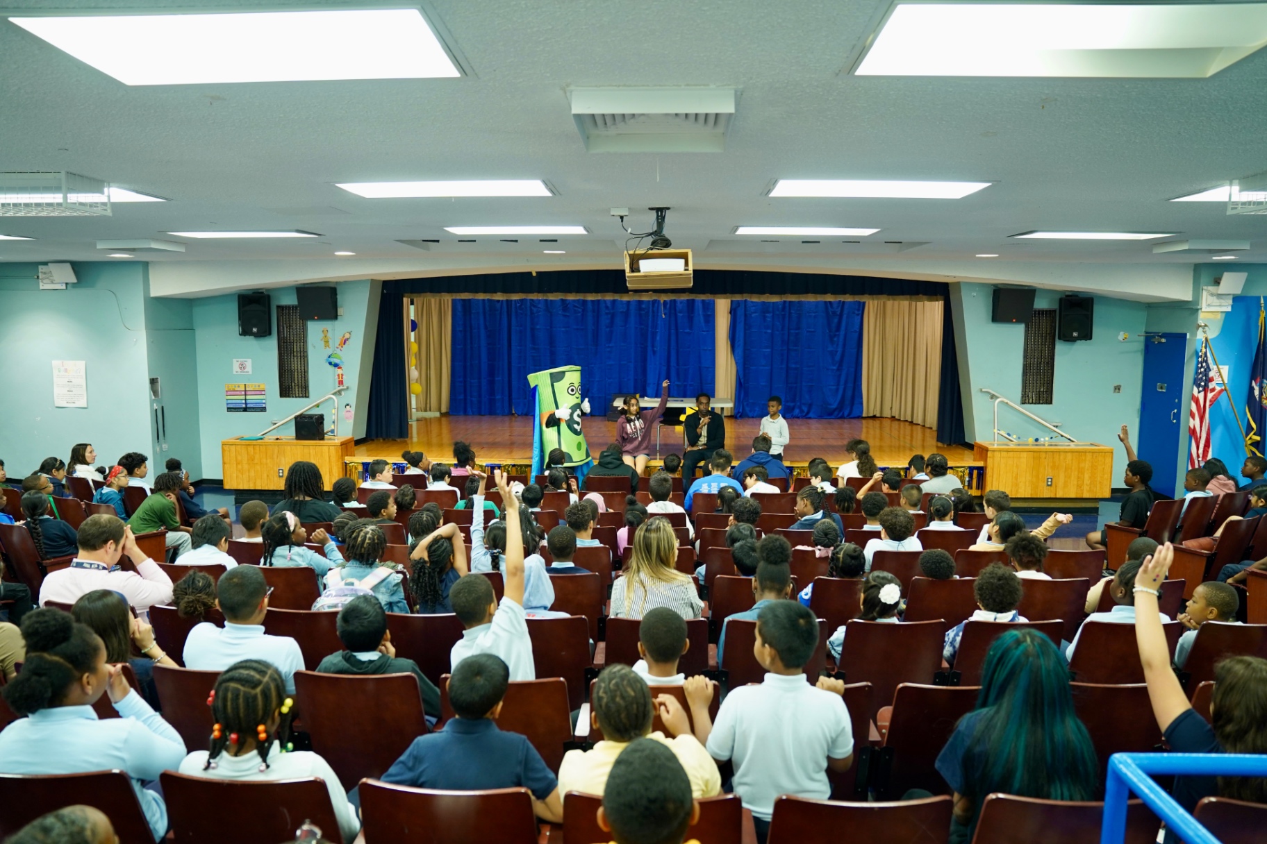 A packed elementary school assembly raising hands during a money lesson